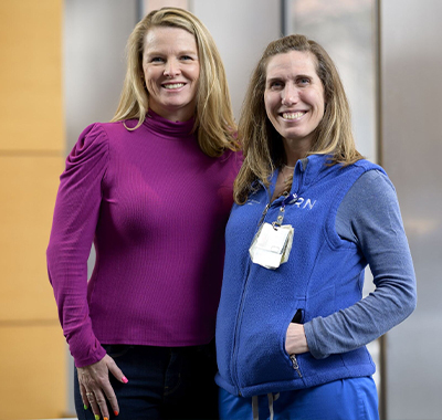 Two women posing for a photo together in front of a wood and silver backdrop. One woman with blonde hair, in a purple turtleneck. The other woman with dirty blonde hair, in a blue vest, with a MSK badge on.