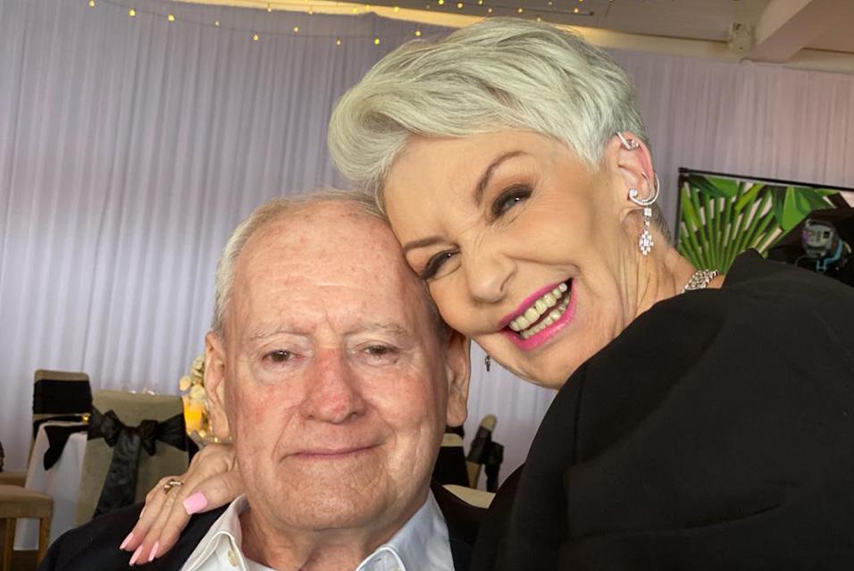 A woman with short white hair smiles at the camera, her head pressed against that of a white-haired man seated next to her. 