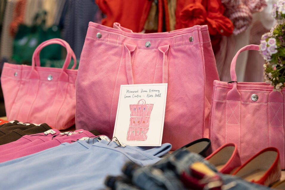 A table displaying pink tote bags and other Alex Mill merchandise. A notecard in front of one of the bags reads “Memorial Sloan Kettering Cancer Center x Alex Mill.”