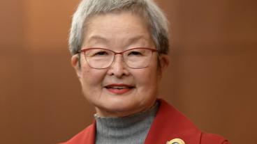 A woman with short gray hair in a gray mock turtleneck sweater and red blazer with a gold brooch stands, smiling, against a wood backdrop.